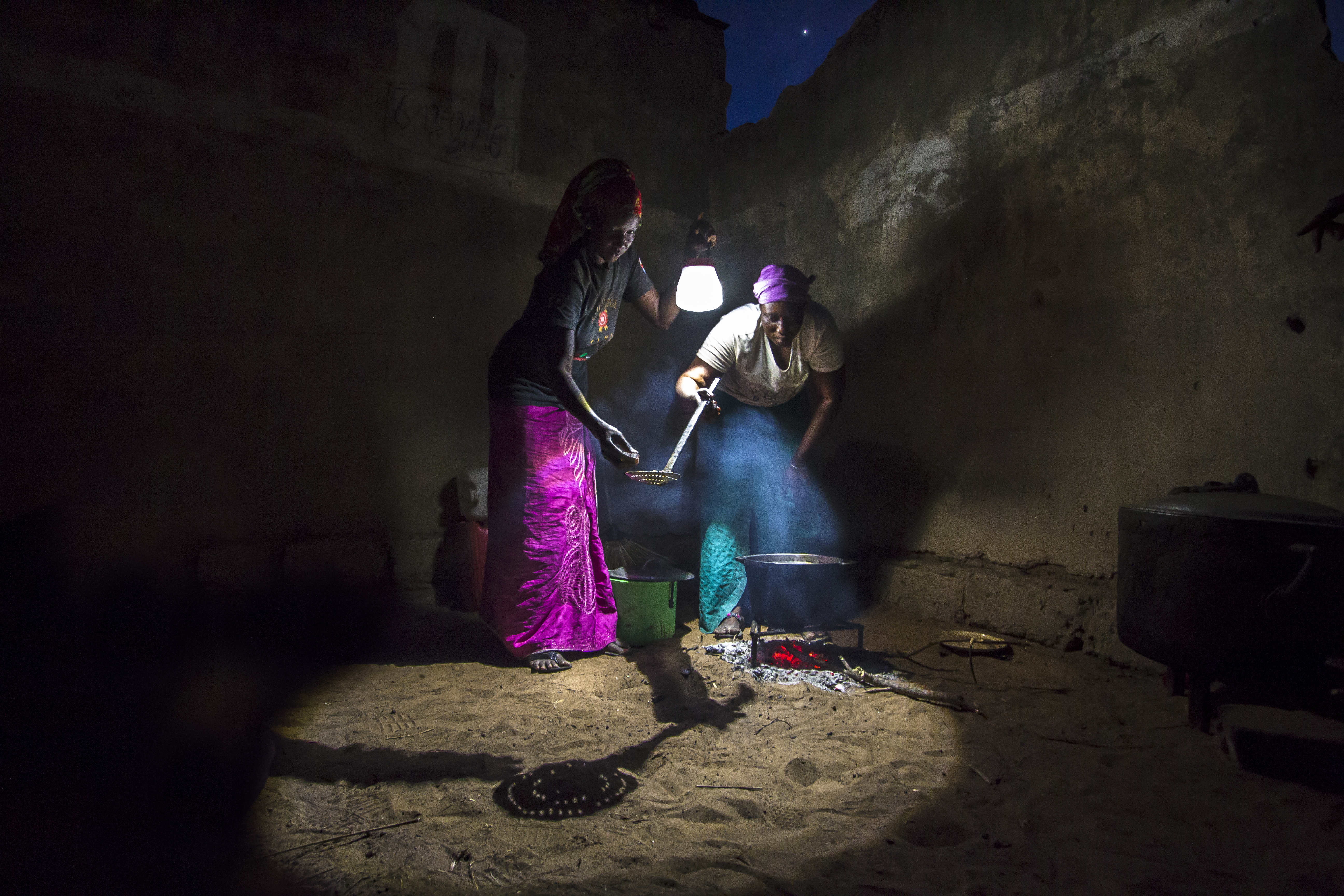 Two women cook over a fire while using BRIGHT Products portable solar lamp SunBell for illumination