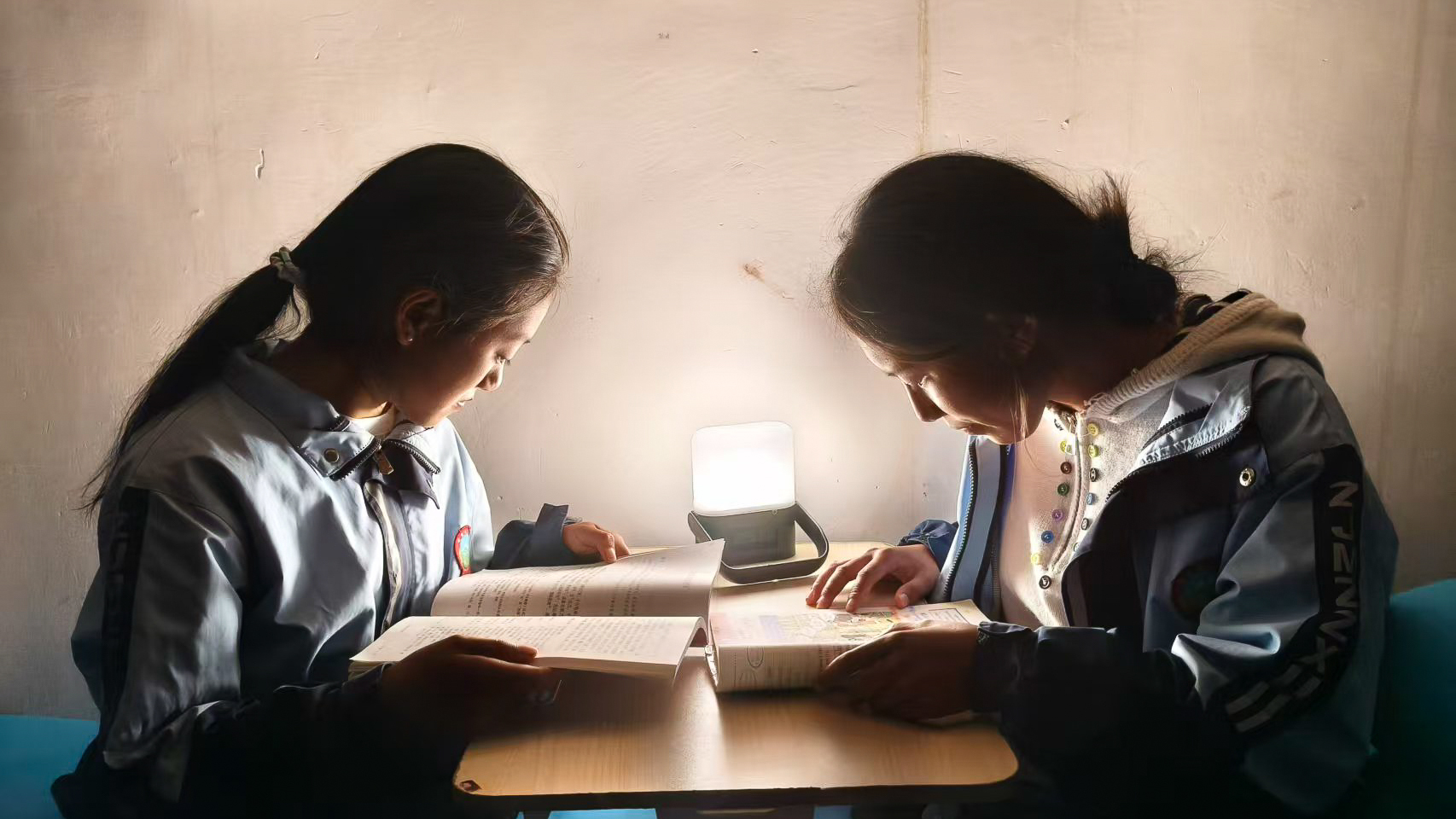 Two students sitting at a desk reading under the light of a Sol solar lamp in a dimly lit room.
