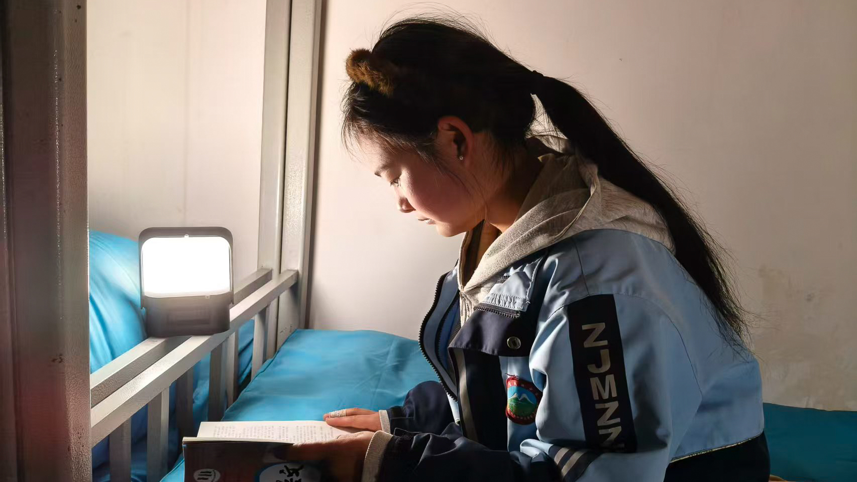 A young student sitting on a bed reading a book under the light of a portable Sol solar lamp in a dim dormitory room.
