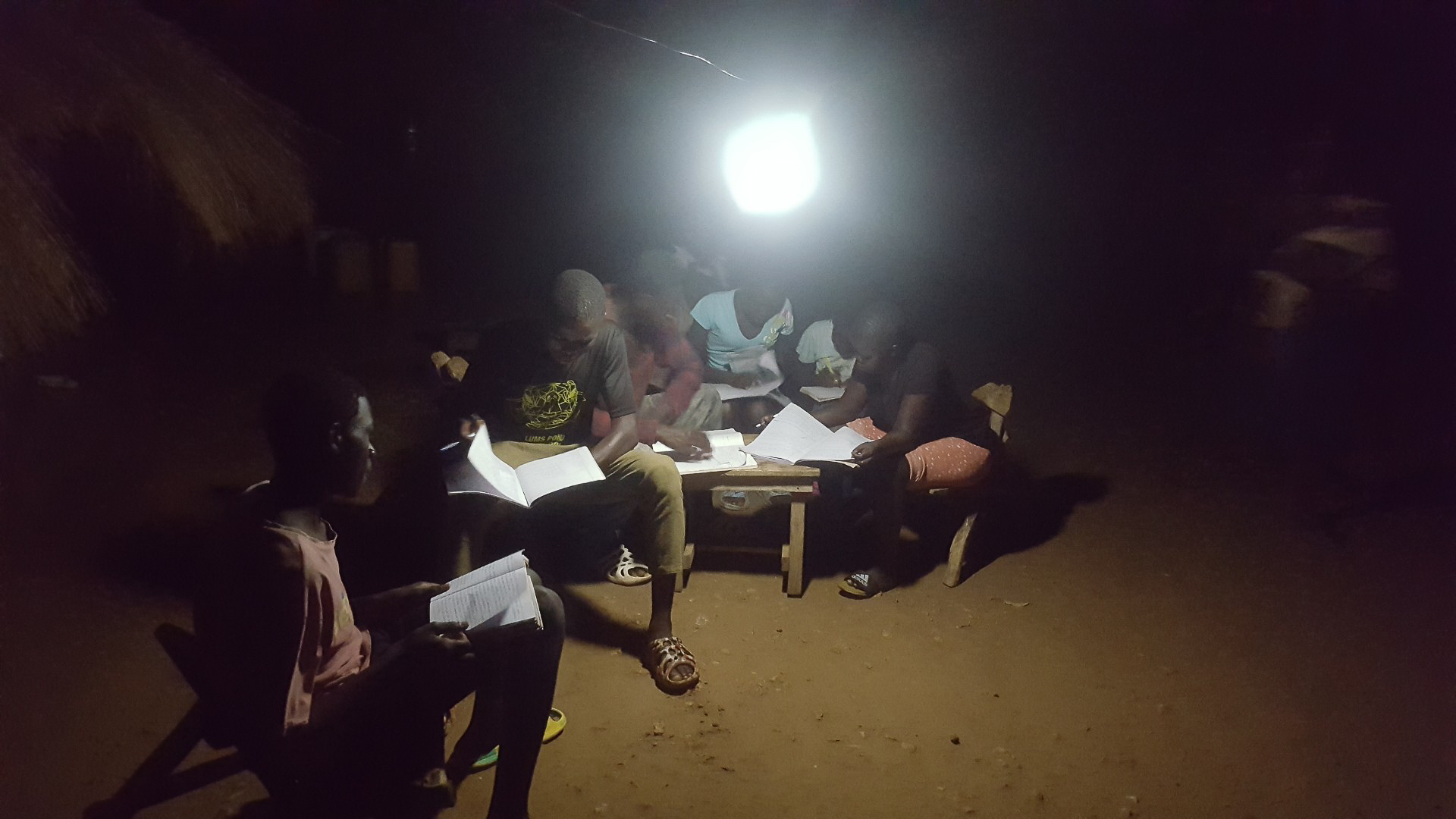 A group of young students gathered at night studying together under the illumination of a portable Sol solar lamp in a refugee settlement.