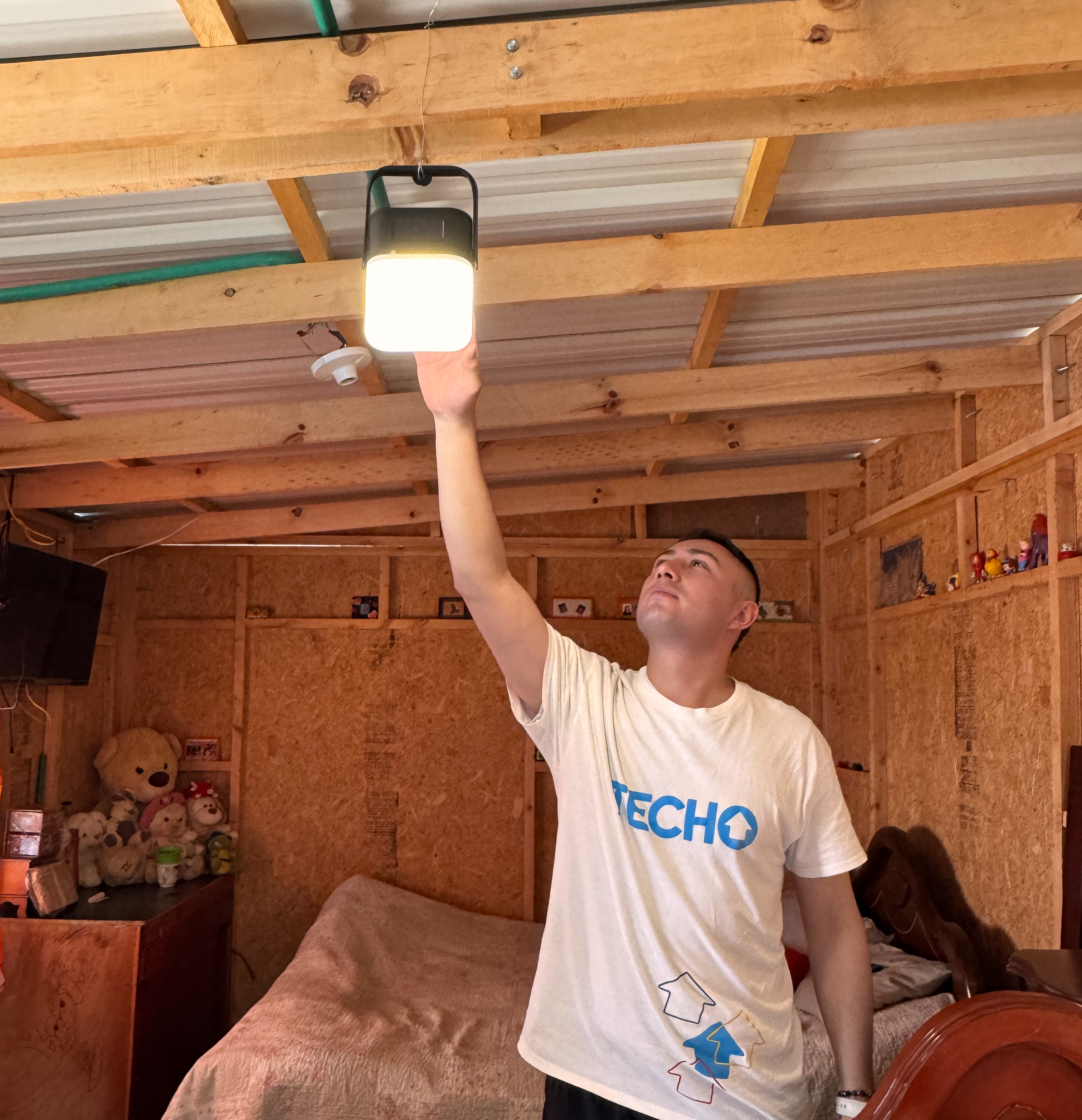 A TECHO representative mounting a Sol Mid Capacity solar lamp on the wall inside a small shelter in an informal settlement.